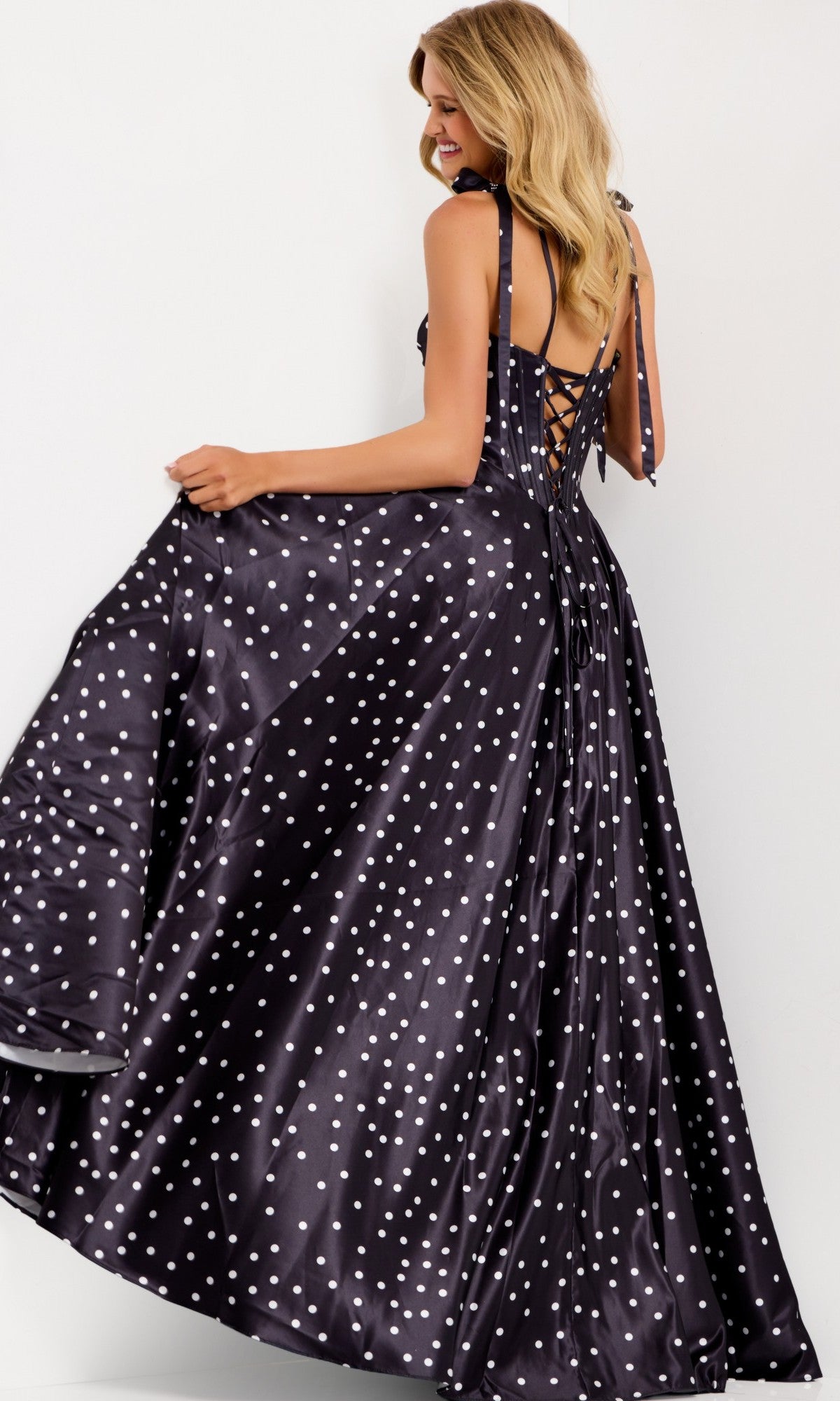 A woman wears the Jovani 49774 long black polka-dot satin prom dress with a corset bodice, lace-up back, and flowing A-line skirt, standing against a plain light background.